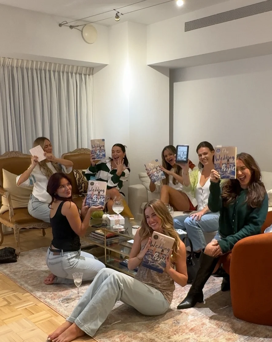 Book club entre amies dans un salon, chacune brandissant son exemplaire de The Wedding People